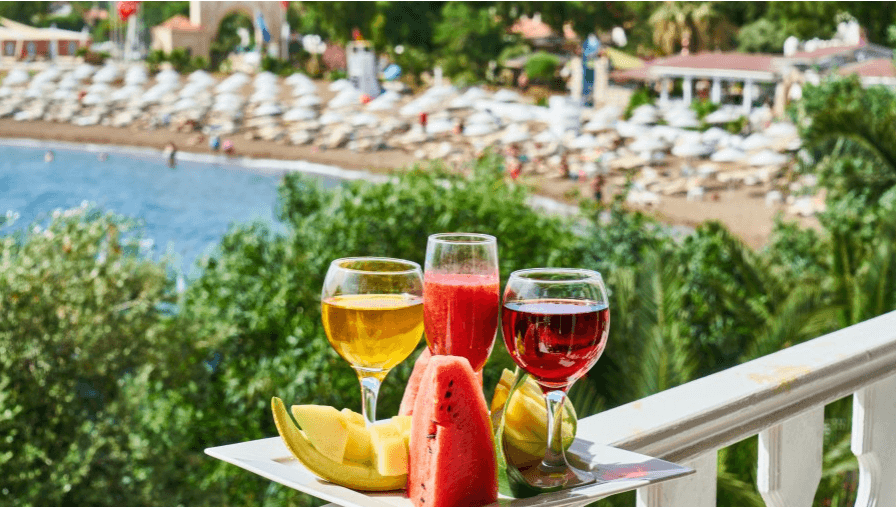 Foto di una serie di bevande di frutta e frutta (ananas e anguria) nappoggiate su una ringhiera, sullo sfondo il mare